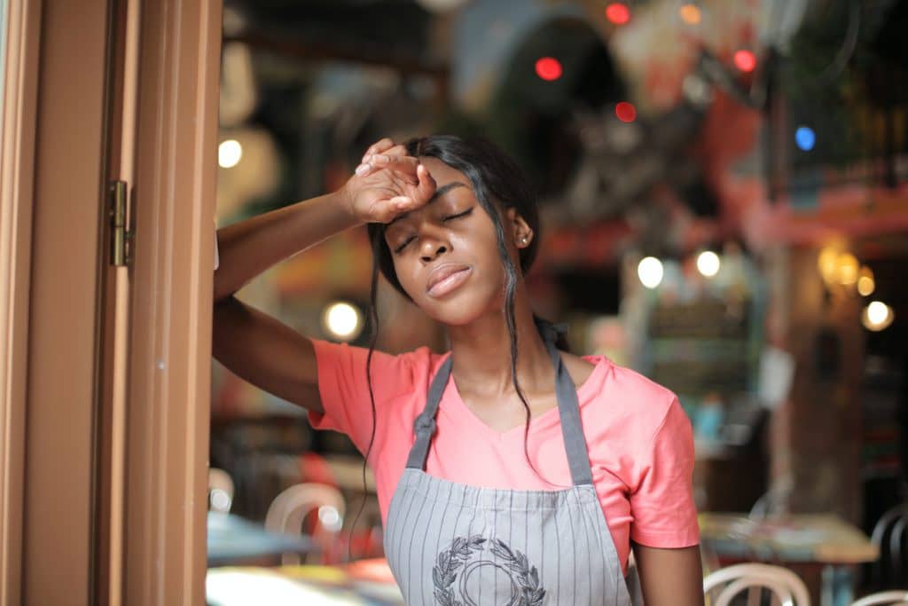 A black girl wearing a pink shirt and a gray apron, leaning on the door, looking tired and dizzy.