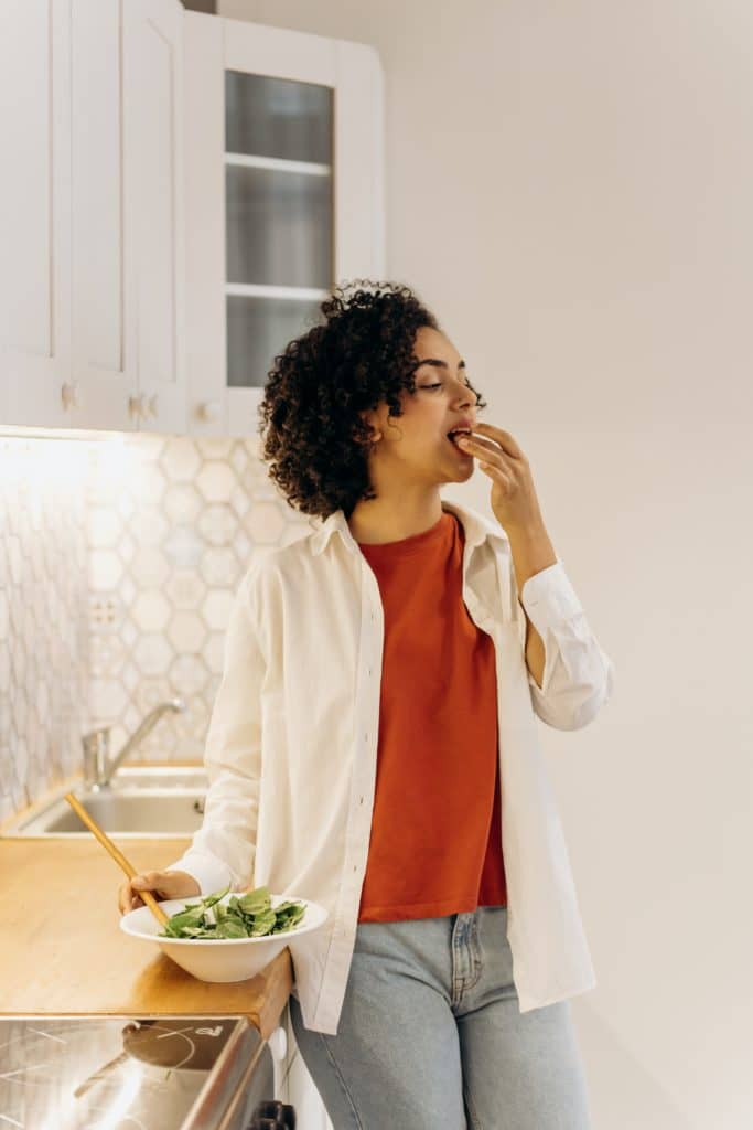 A curly-haired woman in white oversized polo and red shirt eating green vegetables in the kitchen.